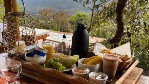 Bandeja de café da manhã servida na varanda, com frutas, pães e a Serra ao fundo