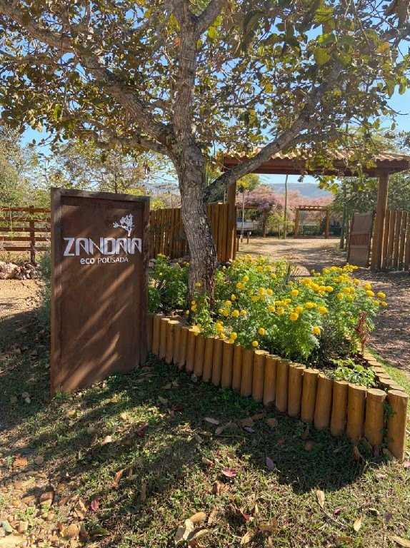 Entrada da Zandaia Eco Pousada, com a placa de madeira, jardim e o portal ao fundo